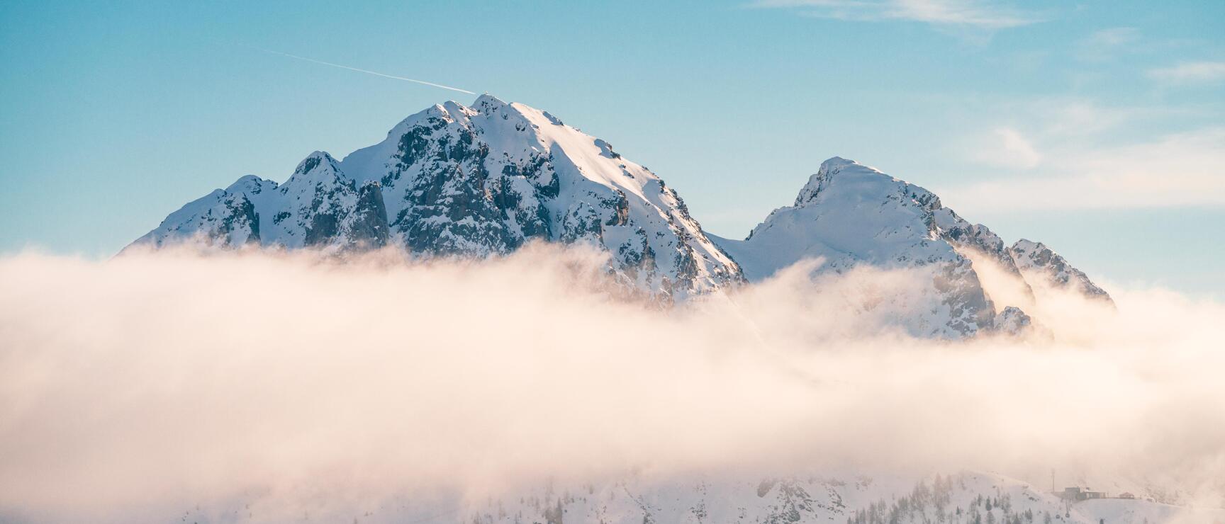 Schneebedeckte Berggipfel ragen über eine sanfte Wolkendecke hinaus, während die klare Winterluft und der strahlend blaue Himmel die alpine Landschaft in ruhiges Licht tauchen.