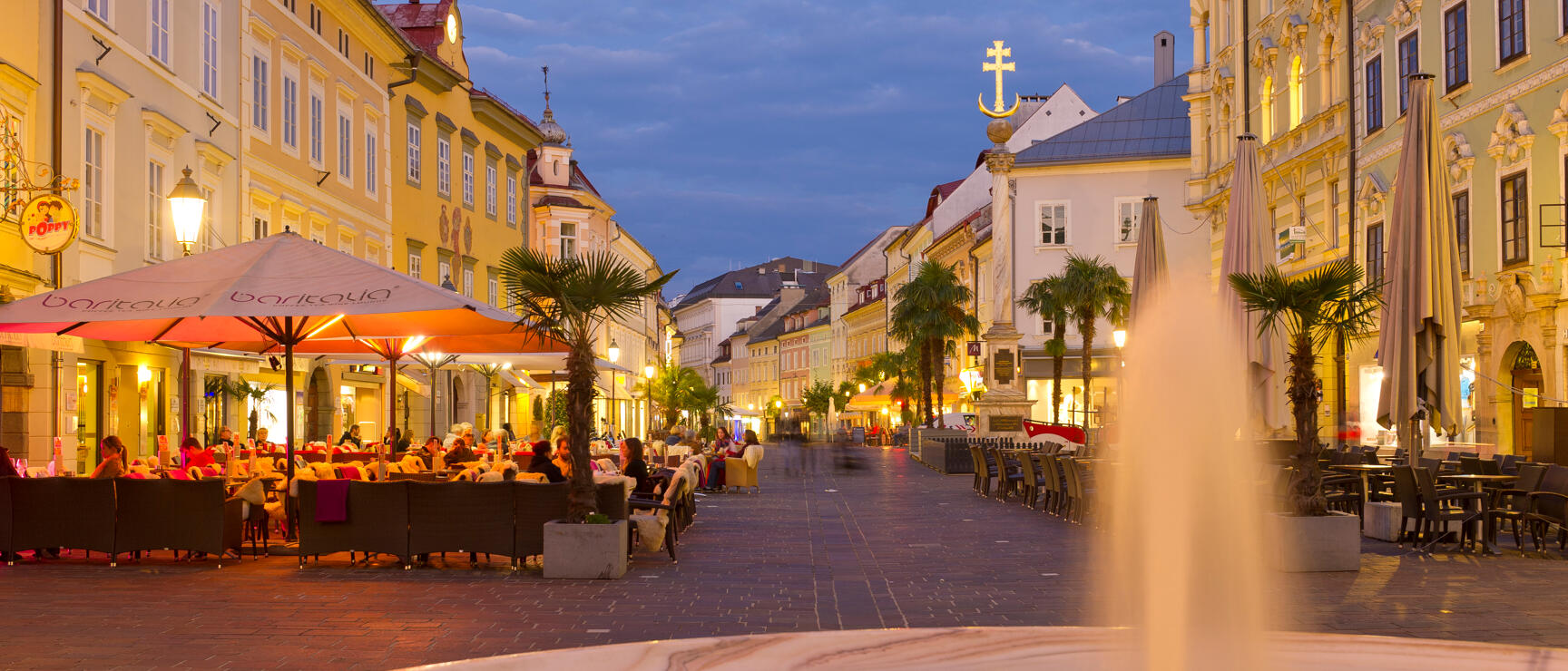 Bustling city square in Klagenfurt at dusk, fountain, palm trees, outdoor seating and illuminated Baroque facades.