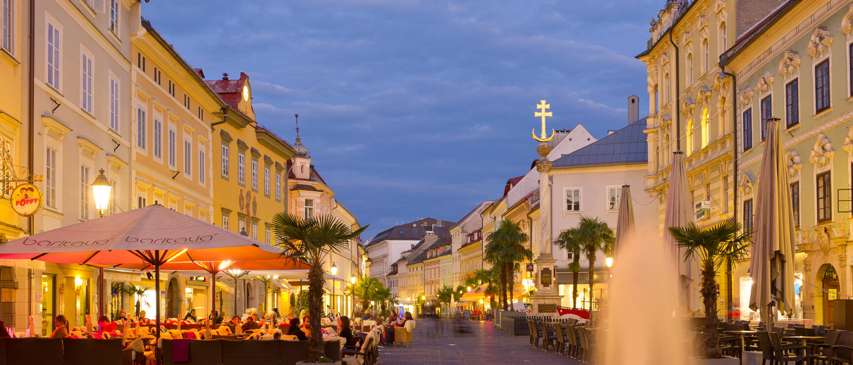 Belebter Stadtplatz in Klagenfurt bei Abenddämmerung, Brunnen, Palmen, Schanigarten und beleuchtete Barockfassaden.