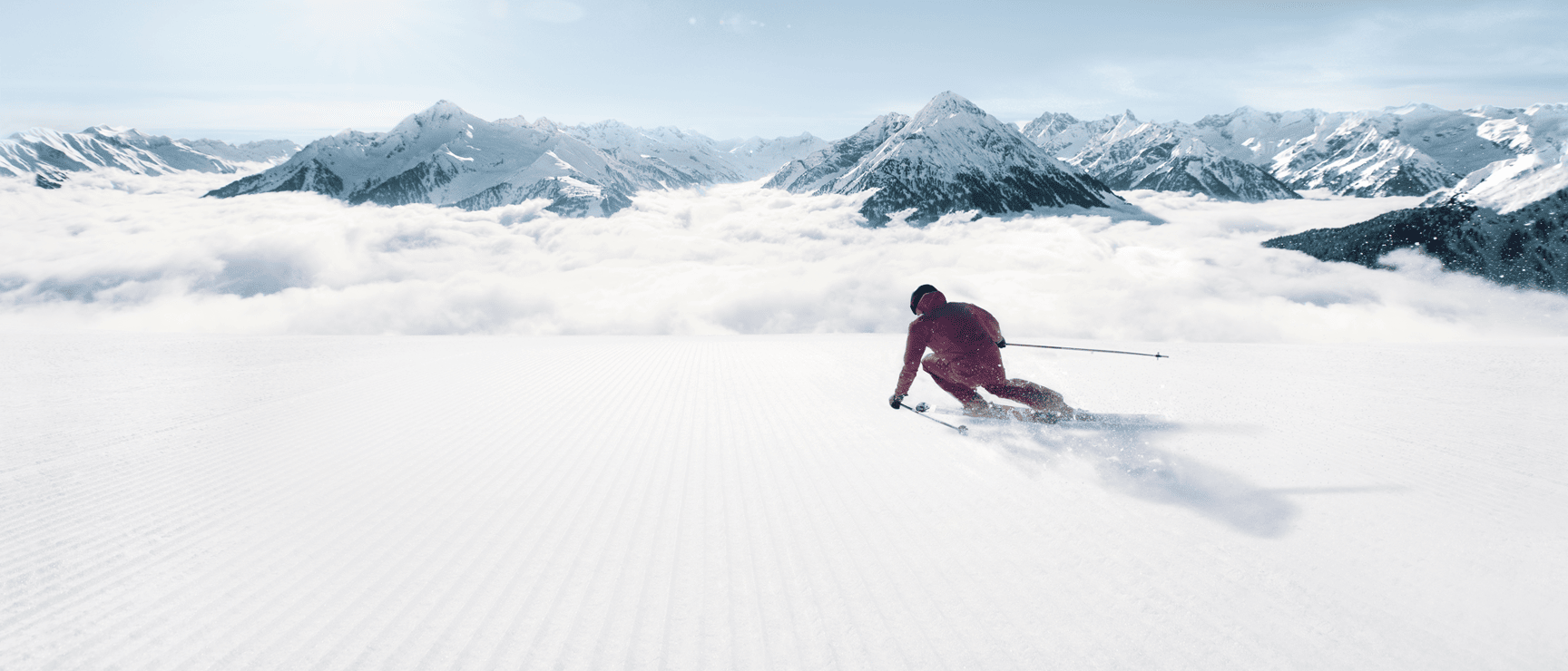 Skier in red jacket on wide slope with snow-covered mountain range and sea of clouds in background.