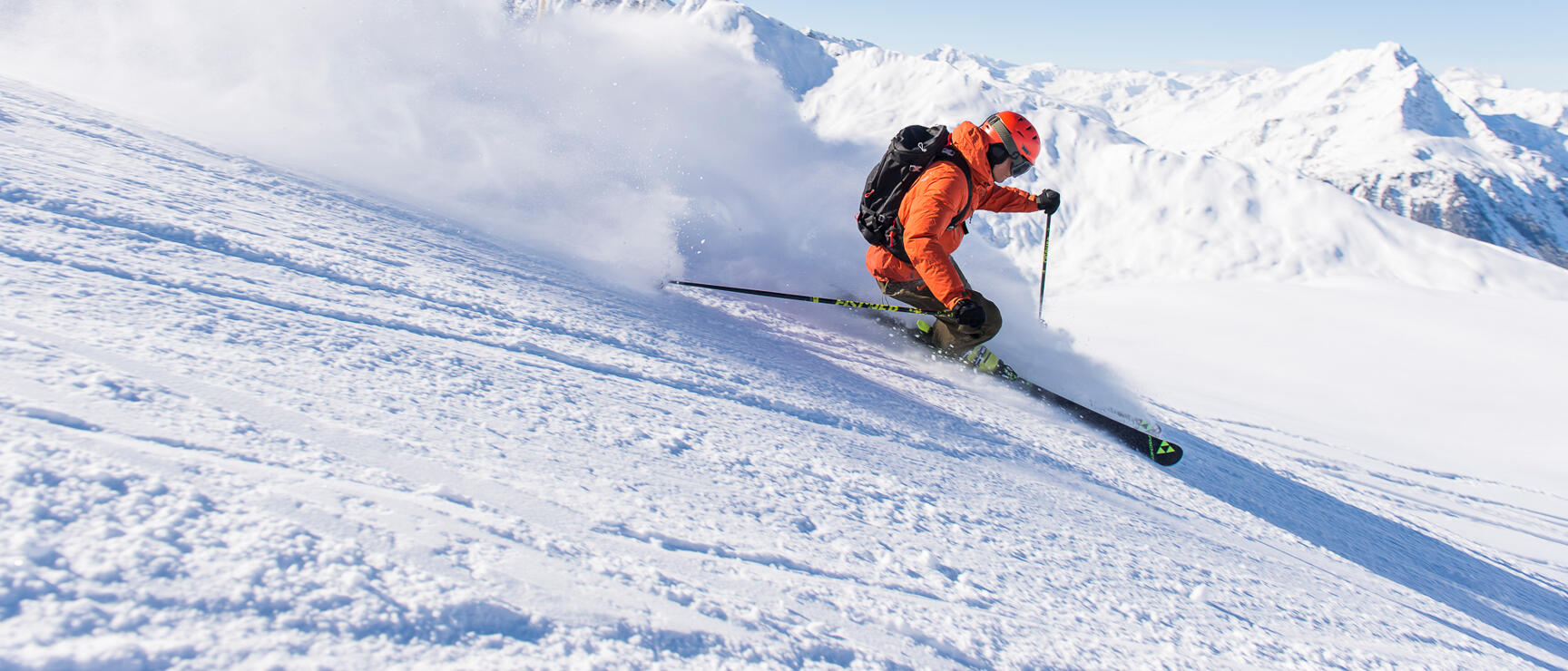 Skier in orange outfit descending steep slope, powder snow spray, snow-covered peaks in background.