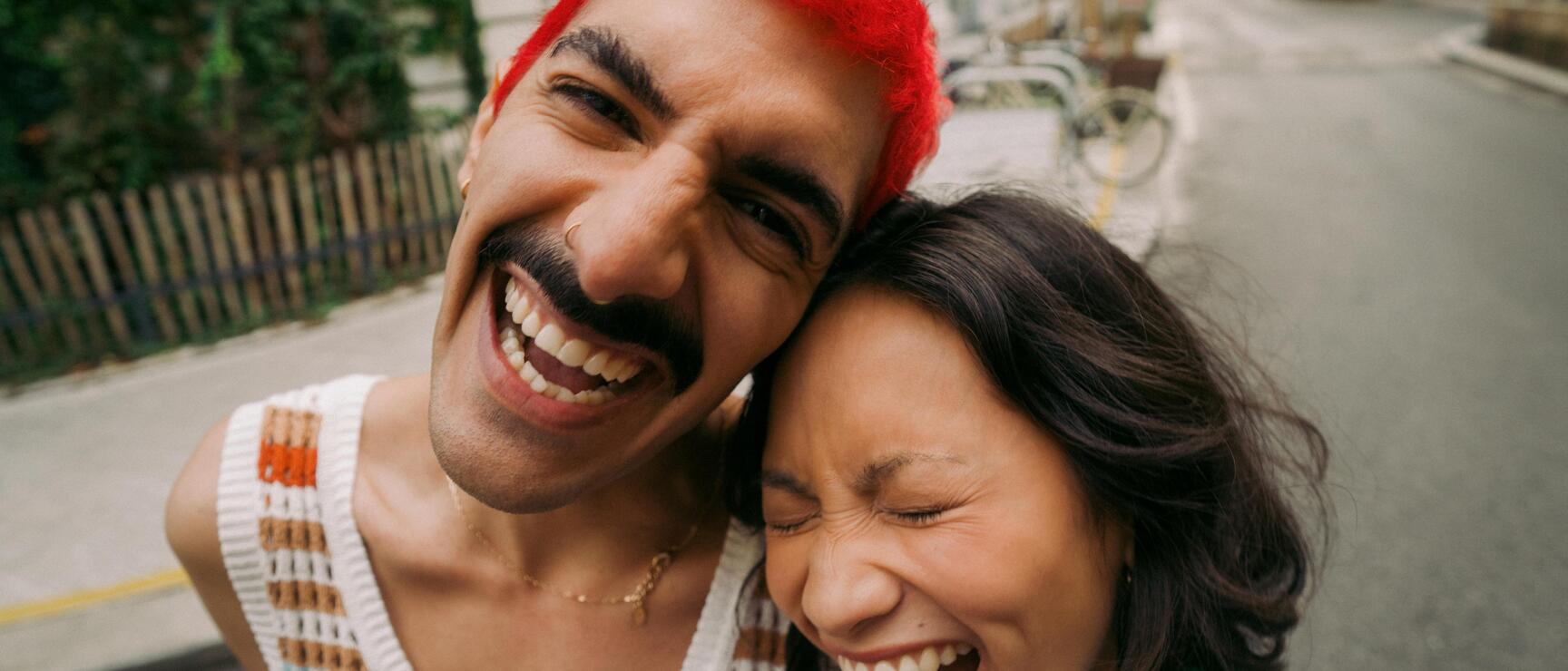 Two laughing people on a city street, man with red hair in tank top, woman in green shirt.