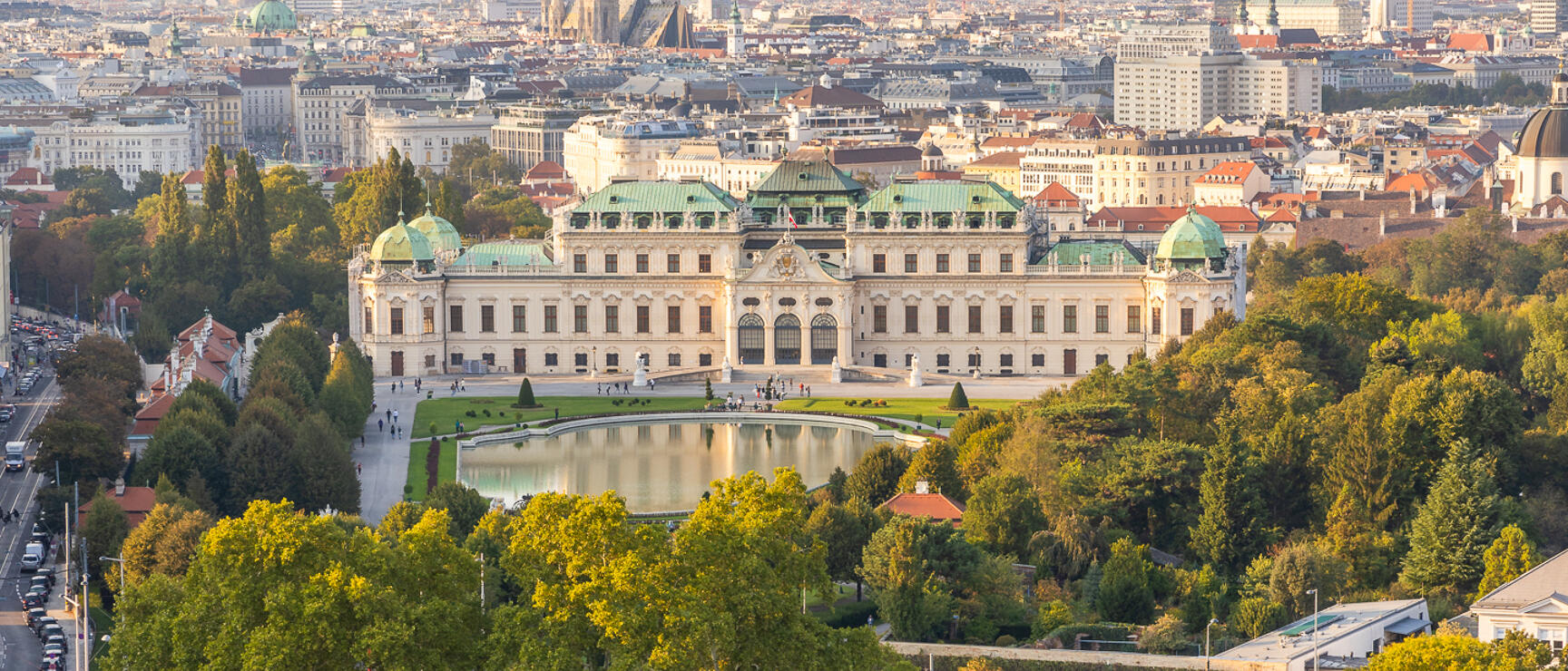 Aerial view of Belvedere Palace with mirror pond, garden landscape and Vienna city skyline in background.