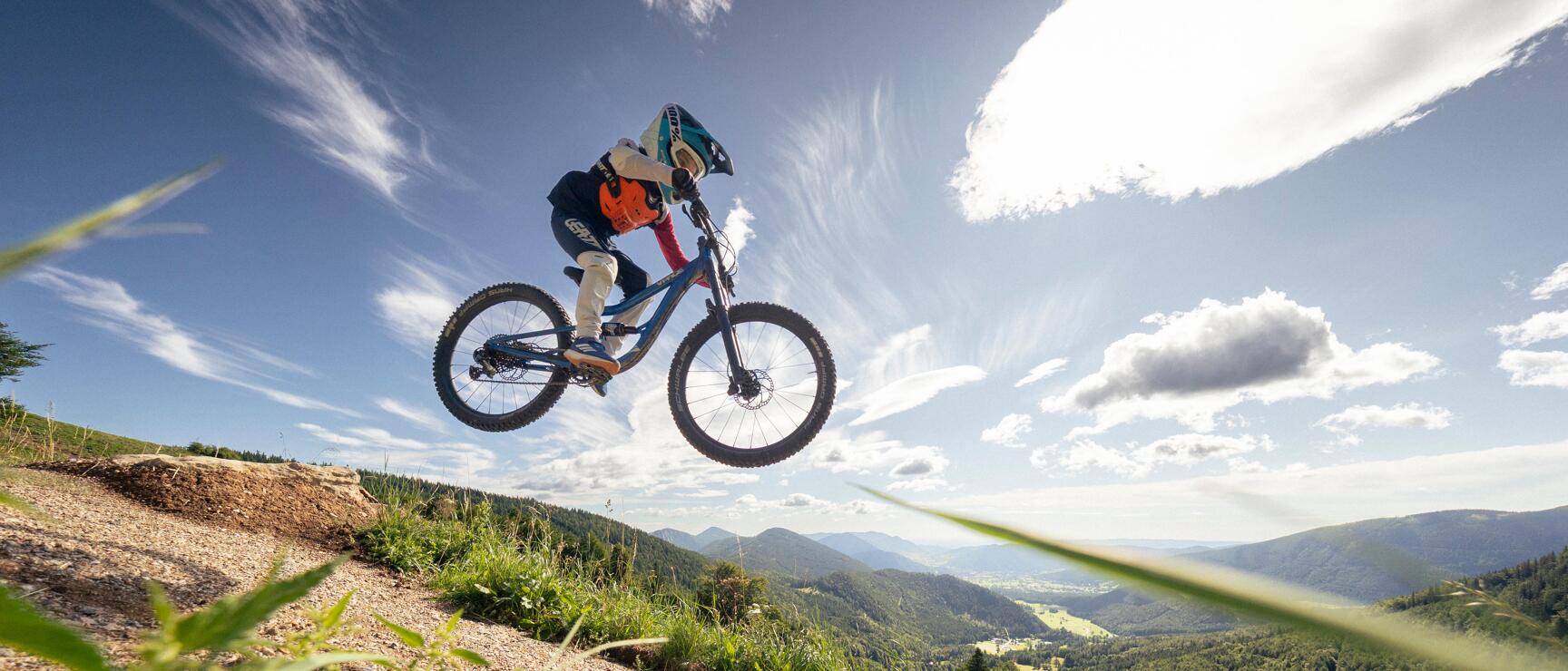Mountain biker launches off a ramp on a bike park trail, green mountain landscape in the background.