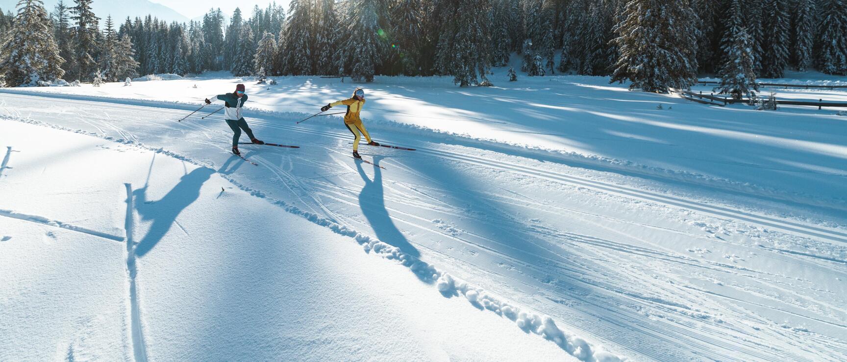 Zwei Personen auf einer Loipe, verschneiter Nadelwald und Berggipfel im Hintergrund, Sonnenschein.