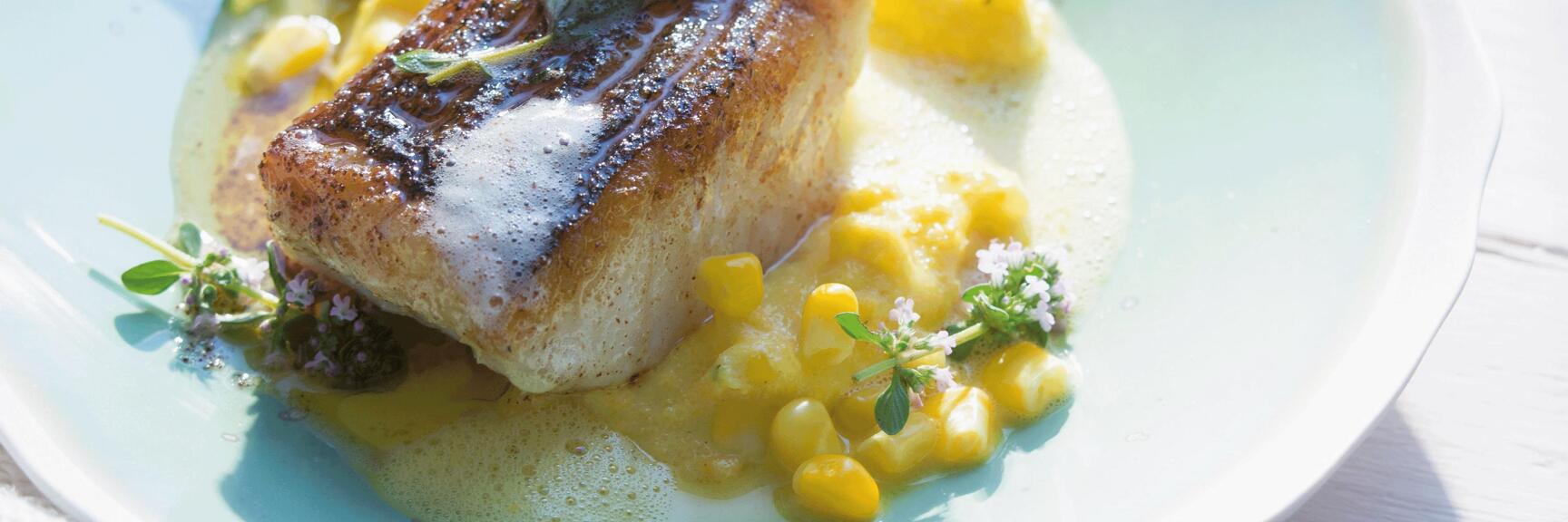 Fried fish fillet with corn kernels and foam on light blue plate, white wooden background.