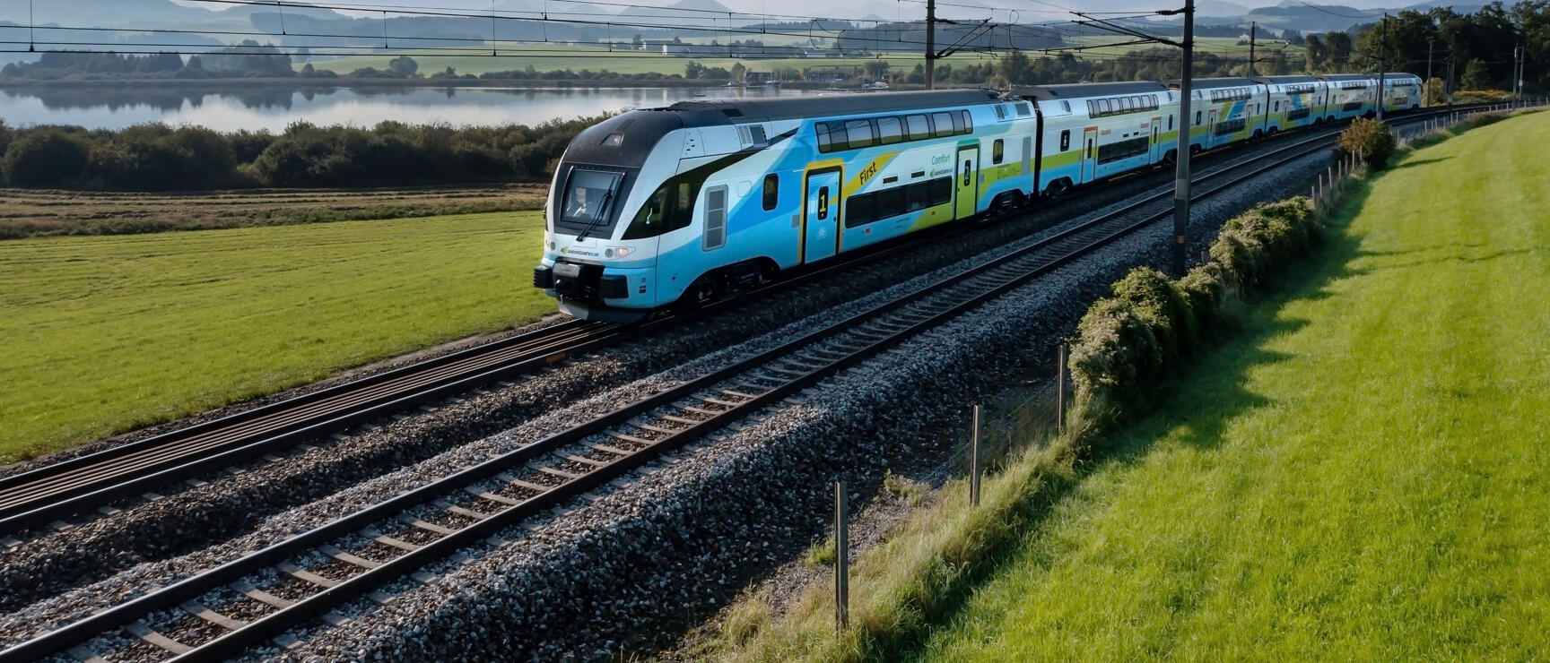 Westbahn train in light blue on tracks, green fields and lake in background, mountains on horizon.