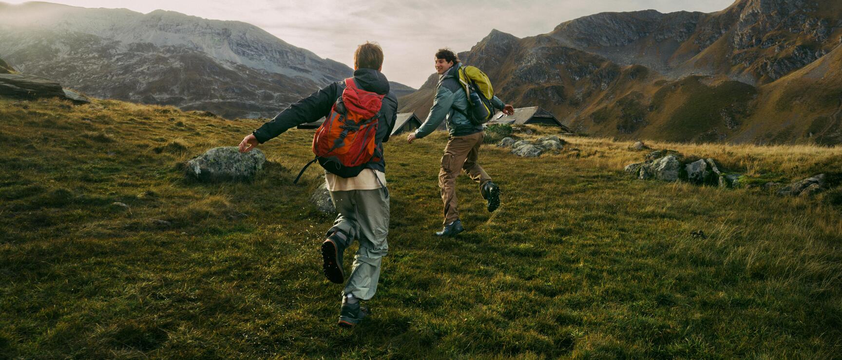 Two hikers with backpacks walking across an alpine meadow with mountain peaks and a cottage in the background