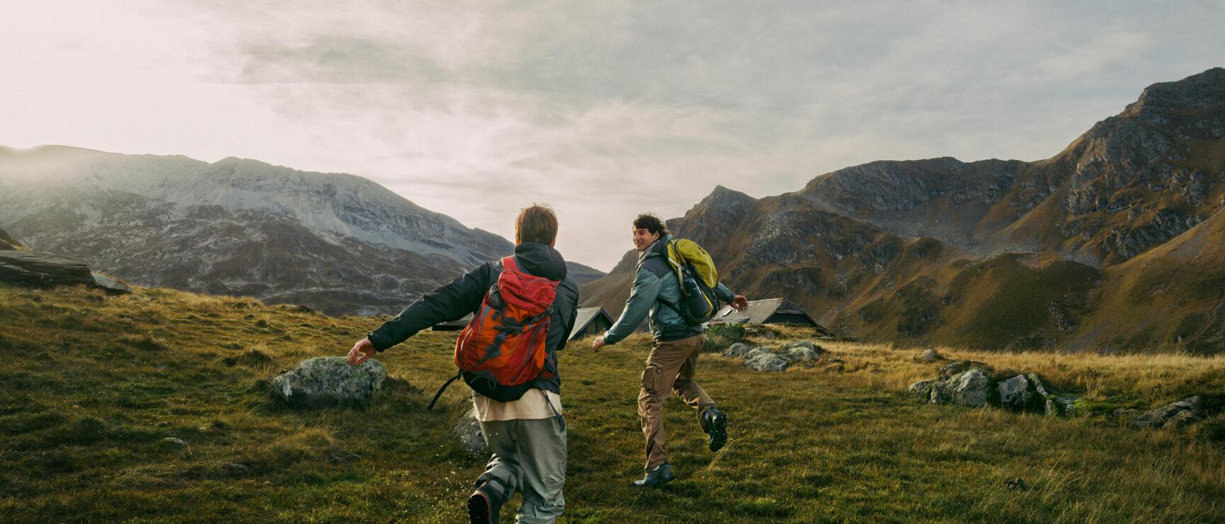 Two hikers with backpacks walking across an alpine meadow with mountain peaks and a cottage in the background