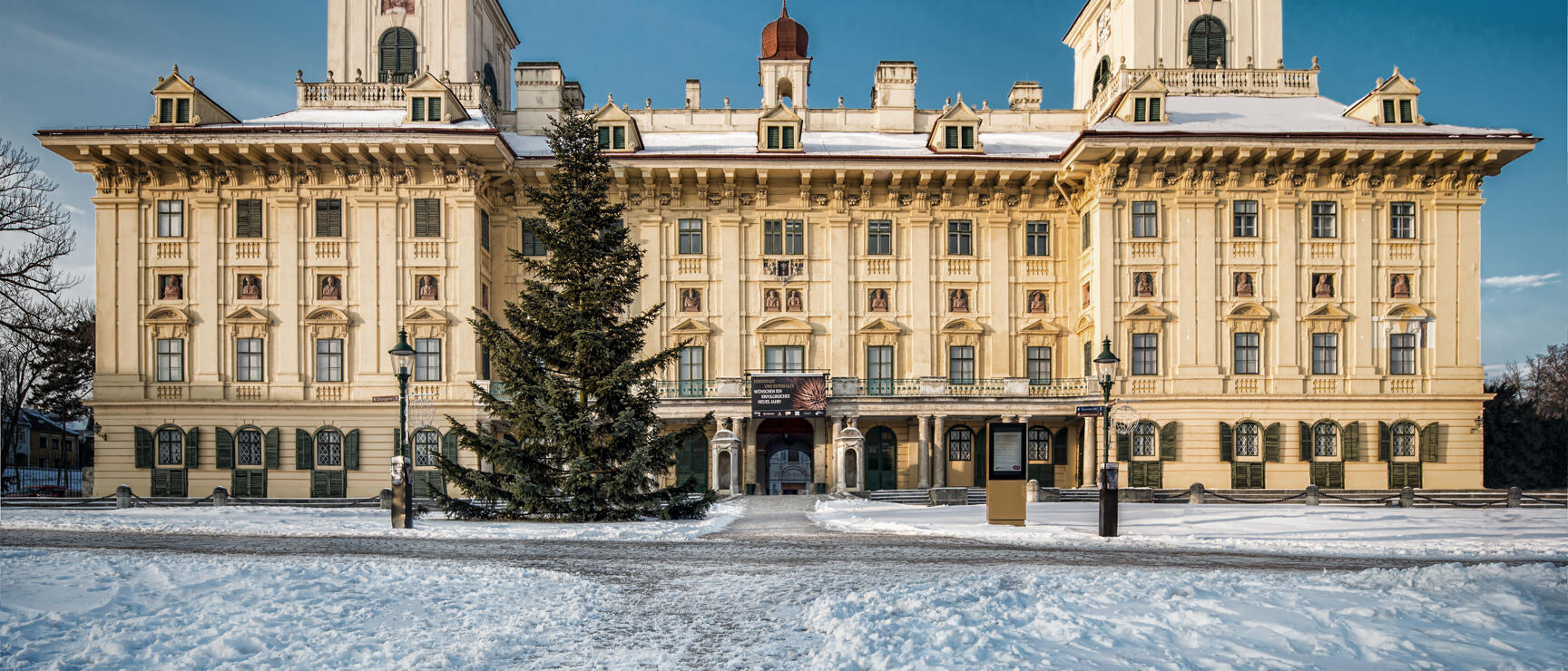 Schloss Esterházy in Eisenstadt im Winter, schneebedeckter Vorplatz mit großem Nadelbaum, blauer Himmel.