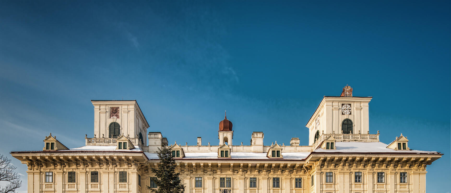 Esterházy Palace in Eisenstadt in winter, snow-covered forecourt with large conifer tree, blue sky.