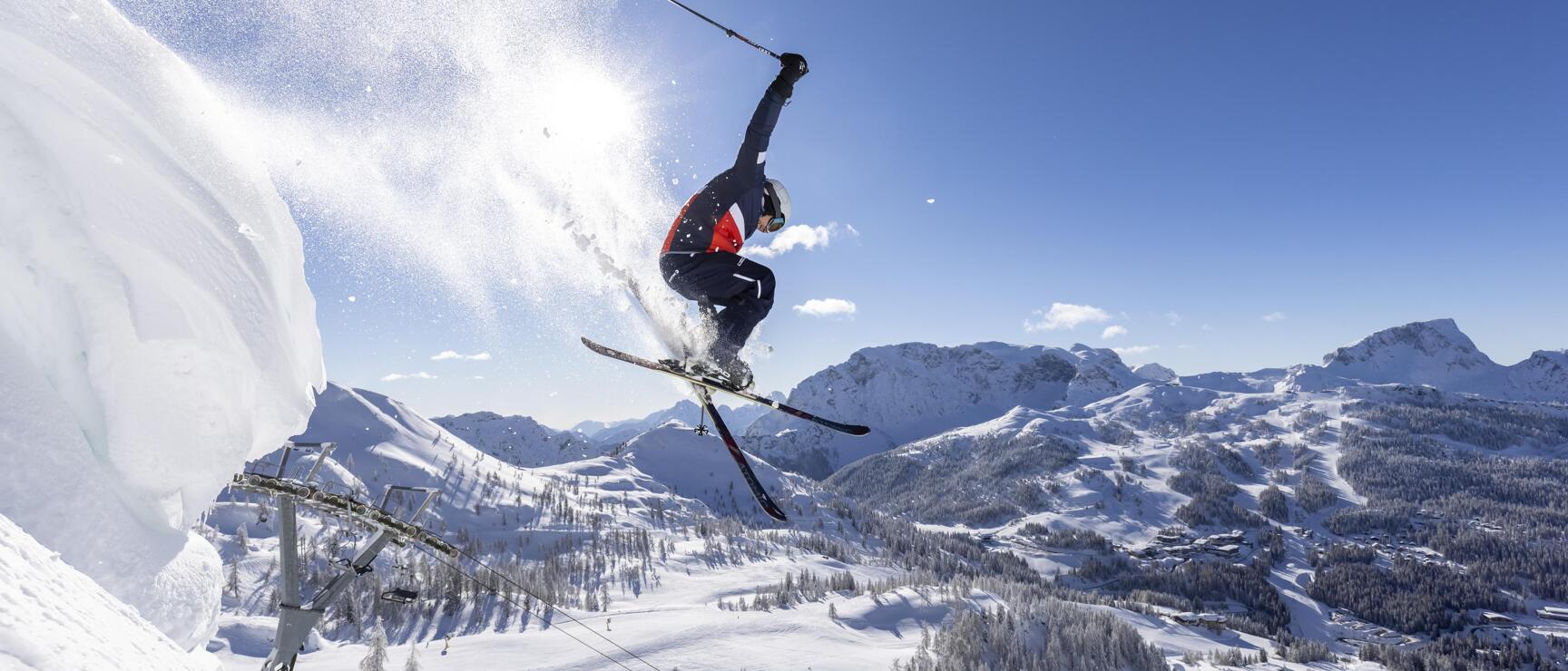 Skifahrer in der Luft bei einem Sprung über eine Schneekante, verschneite Berglandschaft im Hintergrund.