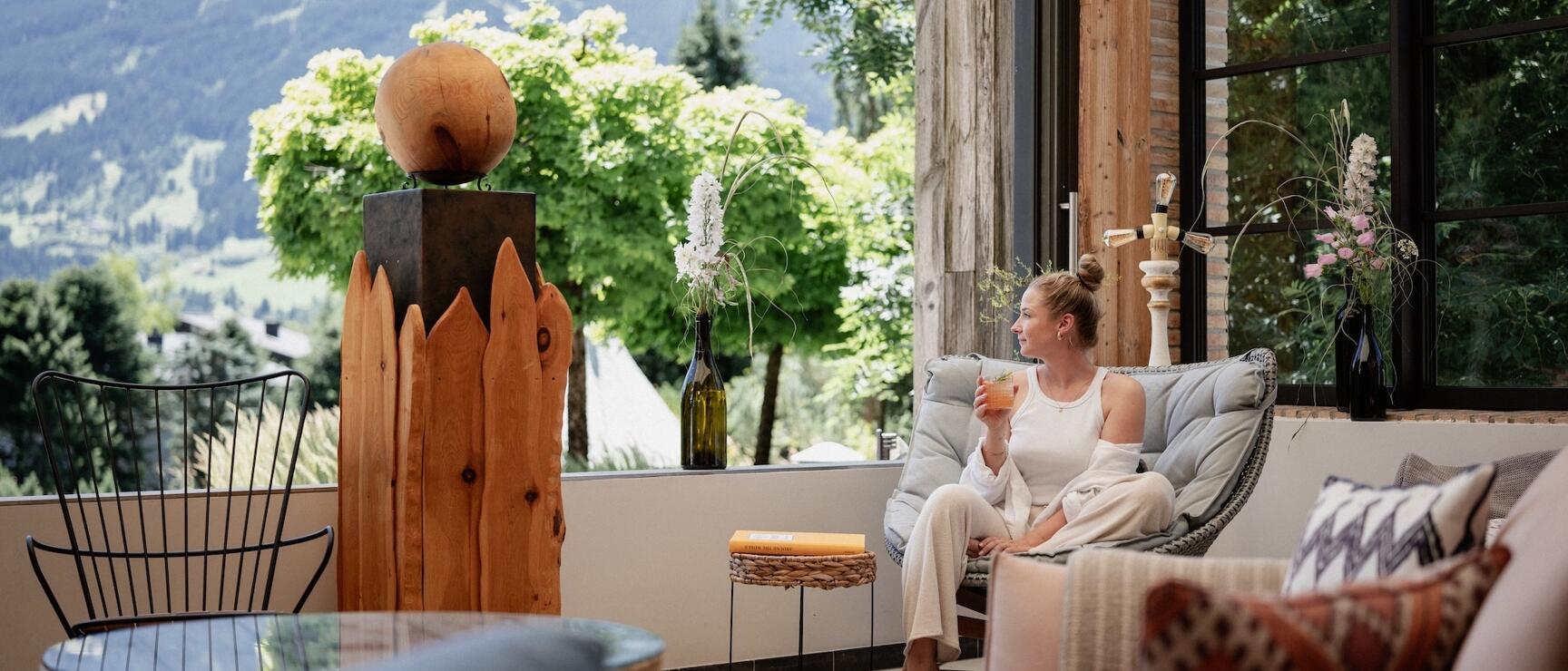 Woman with drink on hotel terrace, wooden sculpture and mountain panorama in background.