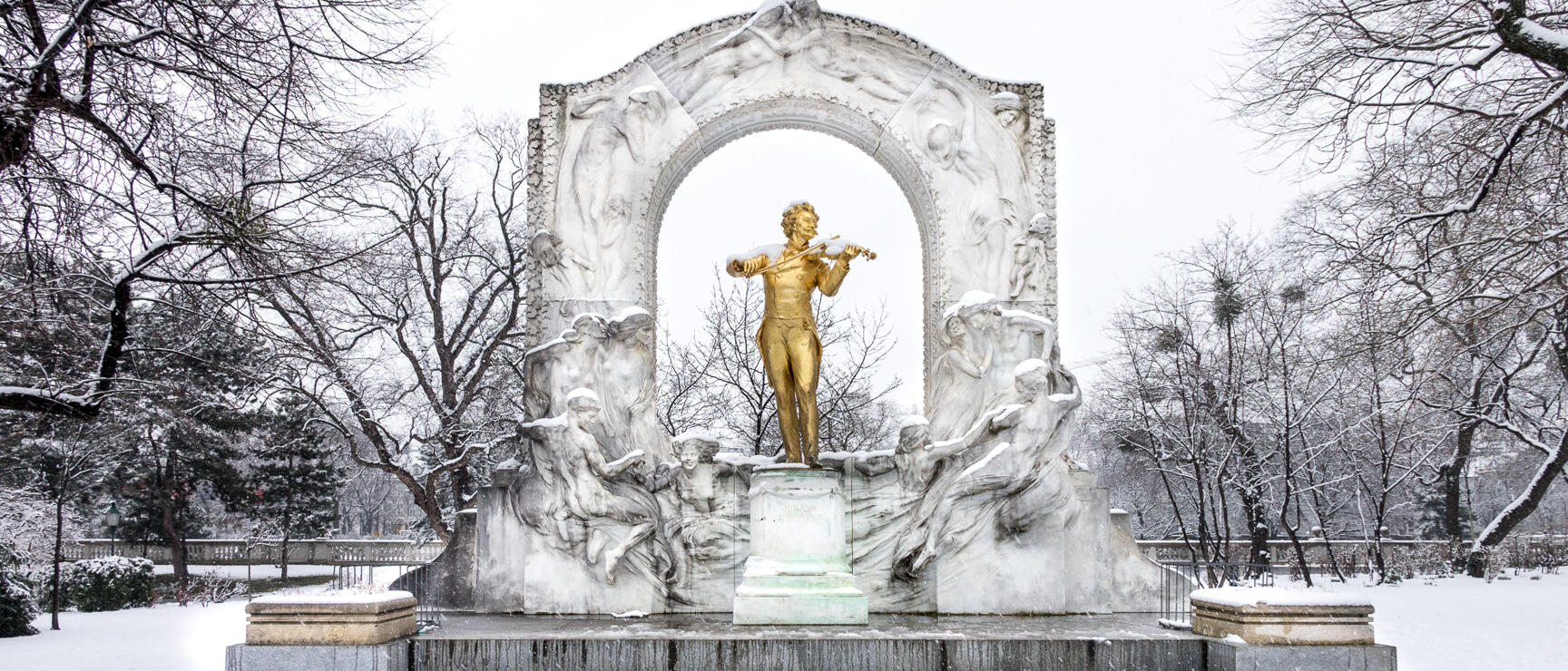 Golden Johann Strauss statue in snowy Vienna city park, white marble arch, bare trees in background.