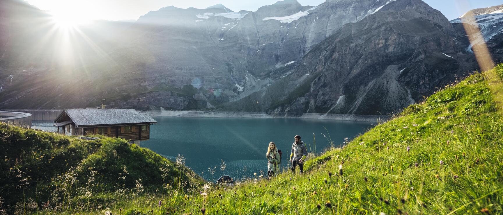Two hikers with backpacks on alpine meadow, mountain lake and wooden hut in the background, glacier and sun star.