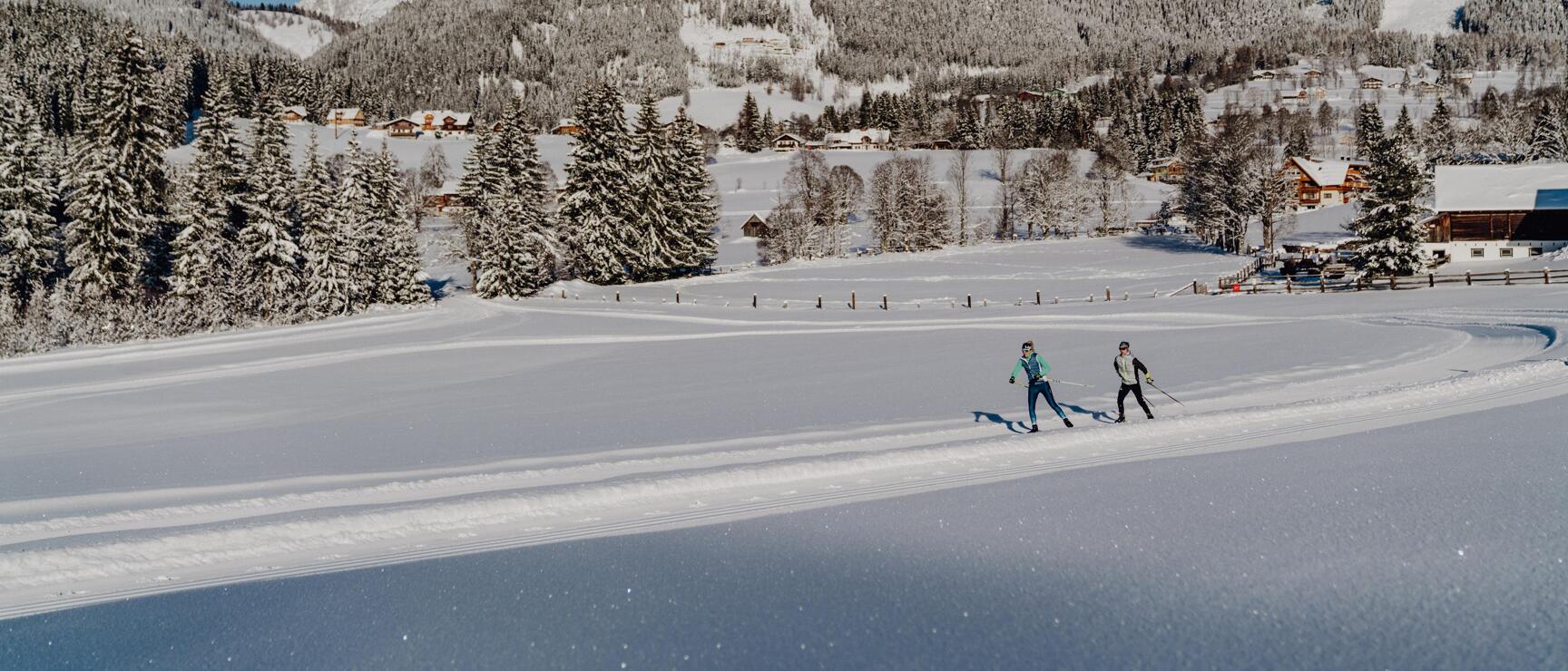 Zwei Personen auf einer Loipe, verschneite Berglandschaft mit Fichten und Gipfeln im Hintergrund.
