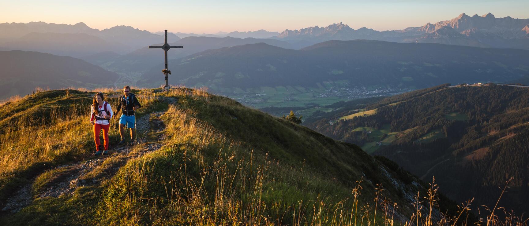 Two hikers beside summit cross with expansive alpine panorama and valley in morning light.