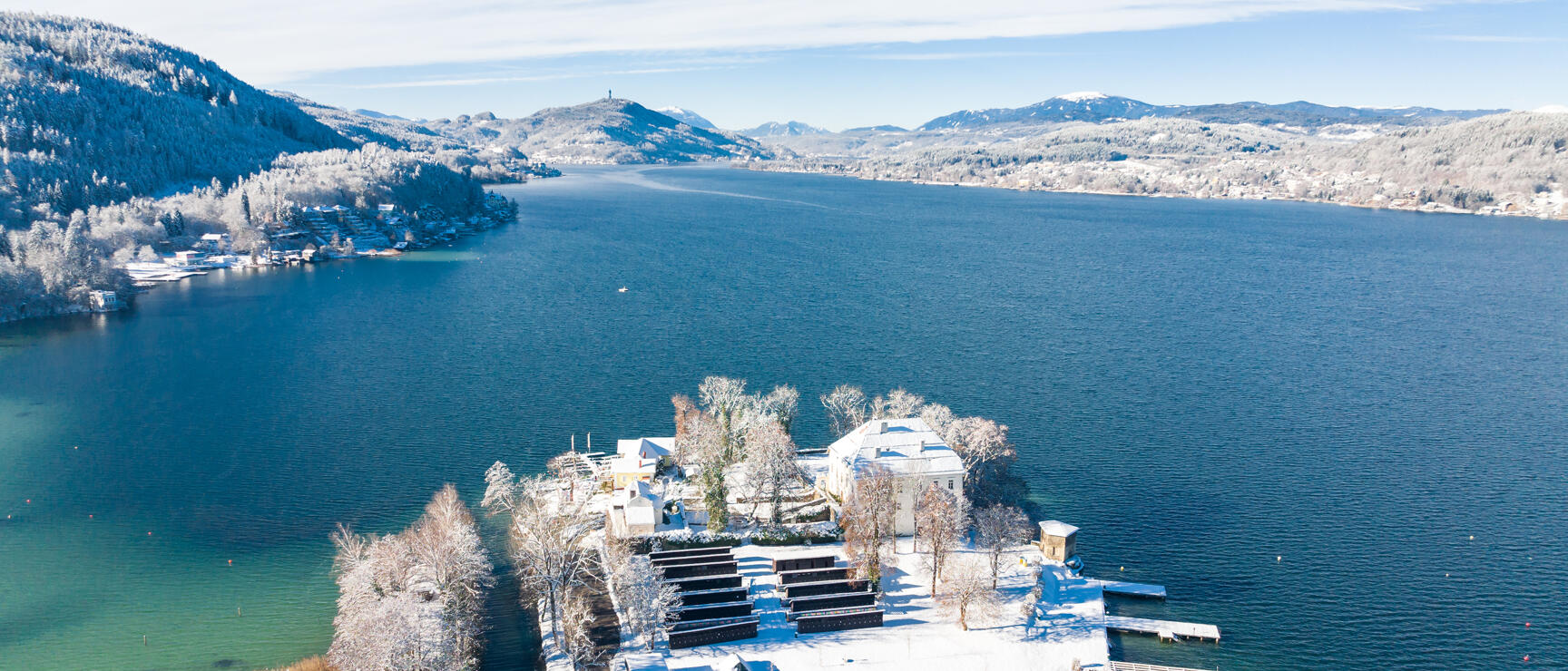 Aerial view of snow-covered lakeside with villa and jetties, blue lake, forested hills in background.