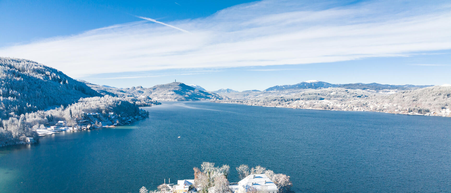 Aerial view of snow-covered lakeside with villa and jetties, blue lake, forested hills in background.
