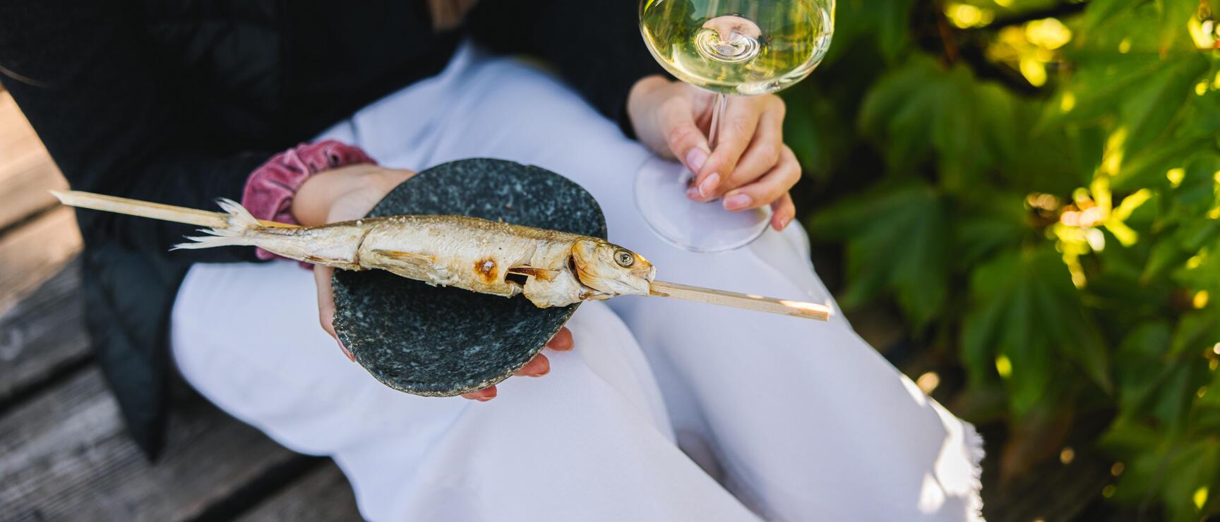 Woman holding grilled fish on skewer on stone and white wine glass, green leaves in background.