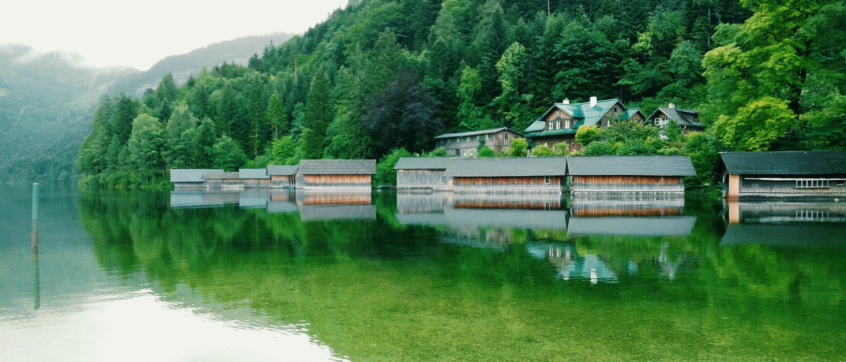 Wooden boathouses at lakeside with reflection in green water, forested hillside and houses in the background.