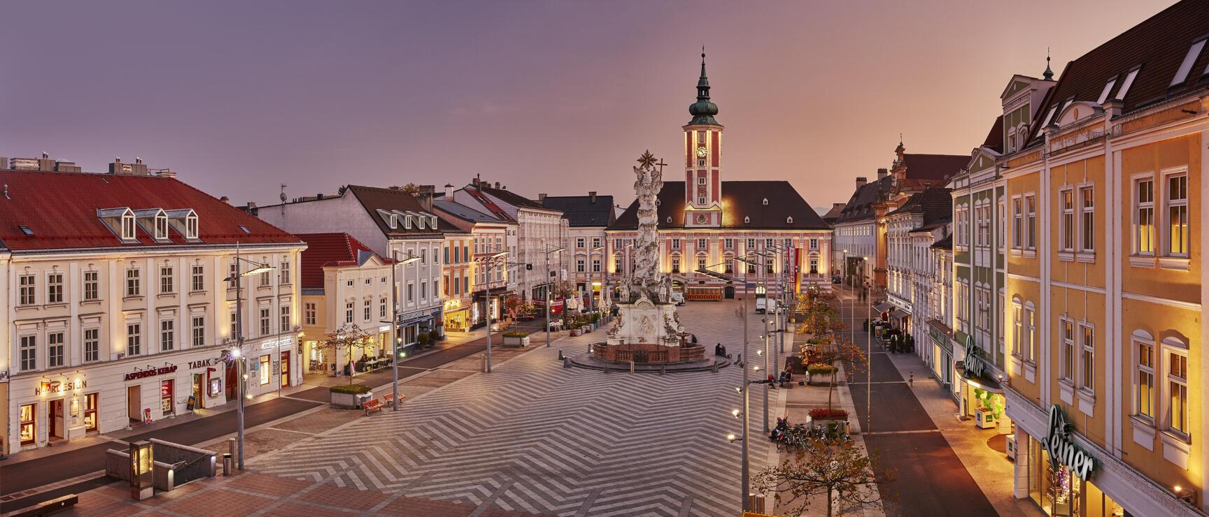 Main square in St. Pölten at dusk with Trinity Column, illuminated town hall and surrounding Baroque facades.