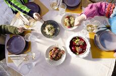 Overhead view of a laid table with multiple dishes, wine glasses and three people dining outdoors.