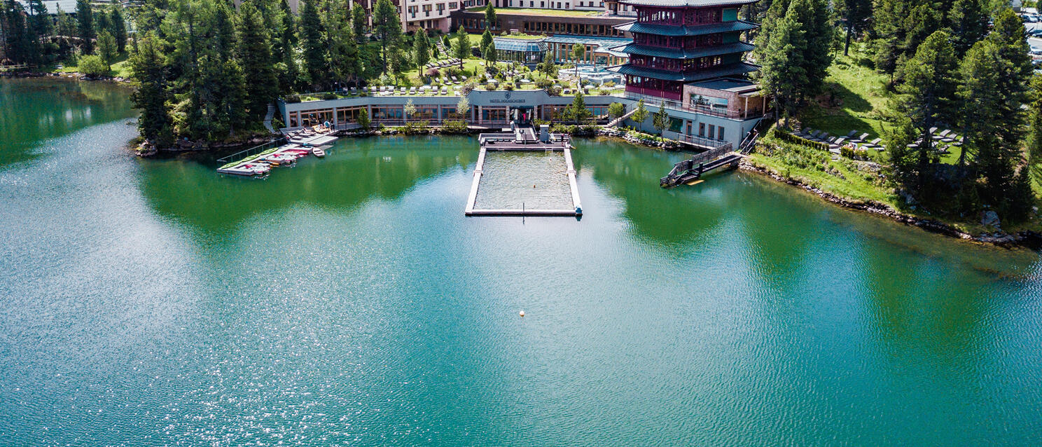 Aerial view of lakeside hotel with pagoda on green mountain lake, coniferous forest in background, bathing dock in water.