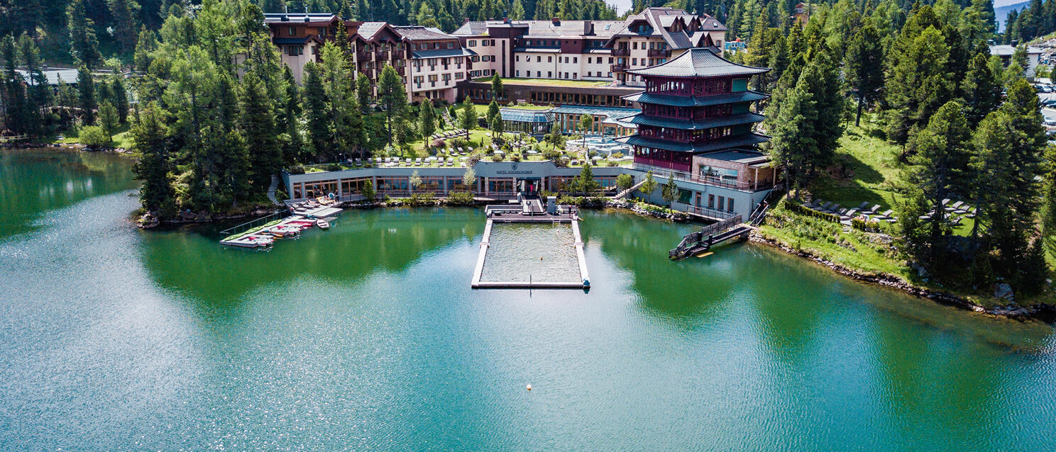 Aerial view of lakeside hotel with pagoda on green mountain lake, coniferous forest in background, bathing dock in water.