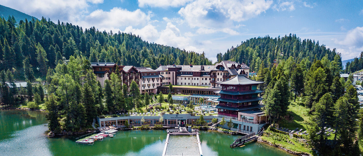 Luftaufnahme eines Seehotels mit Pagode am grünen Bergsee, Nadelwald im Hintergrund, Badesteg im Wasser.