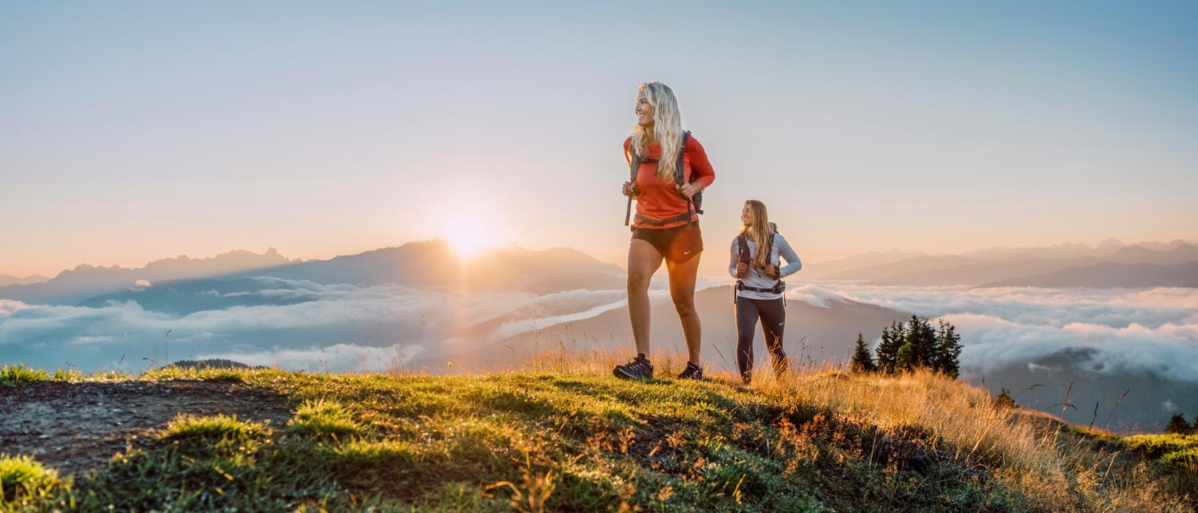 Two female hikers with backpacks on a mountain summit at sunset with sea of clouds and Alpine panorama in the background.