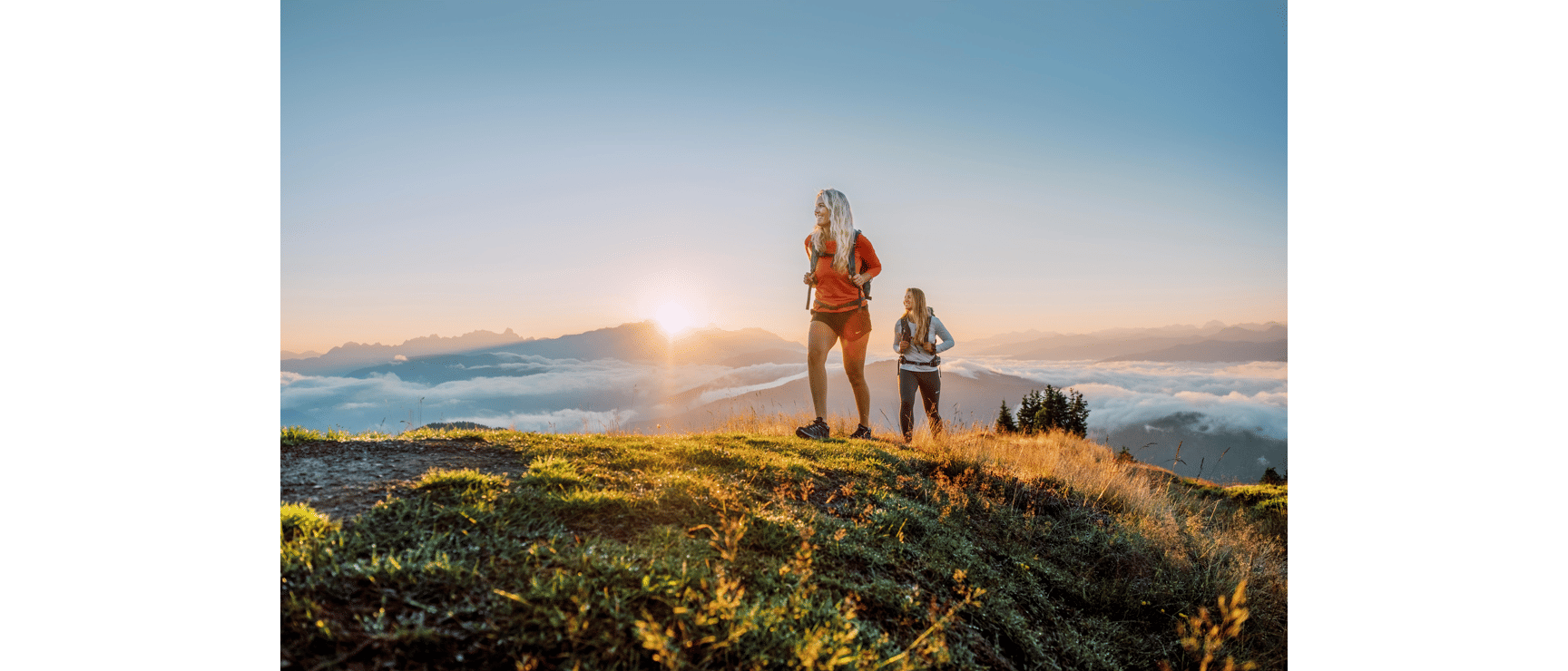 Two female hikers with backpacks on a mountain summit at sunset with sea of clouds and Alpine panorama in the background.