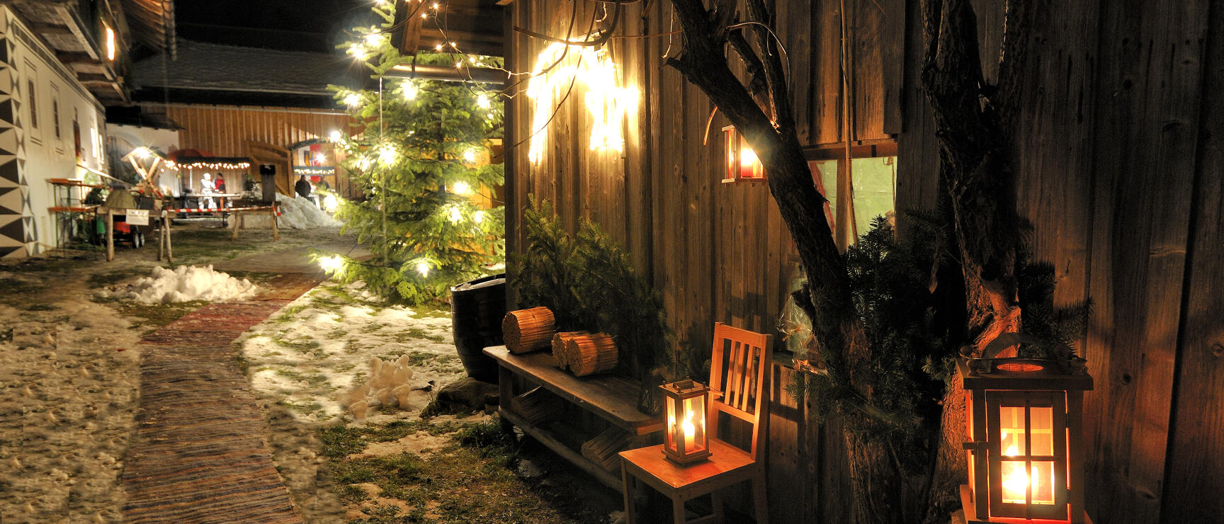 Adventmarkt bei Nacht: beleuchteter Christbaum, Laternen vor Holzscheune, Schnee am Boden, Lichterketten.