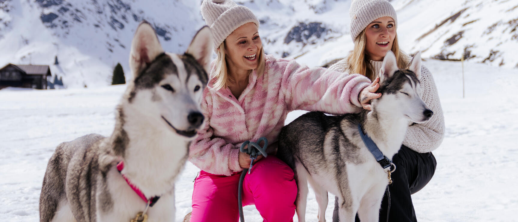 Two women with huskies in snow, snow-covered mountains in background.