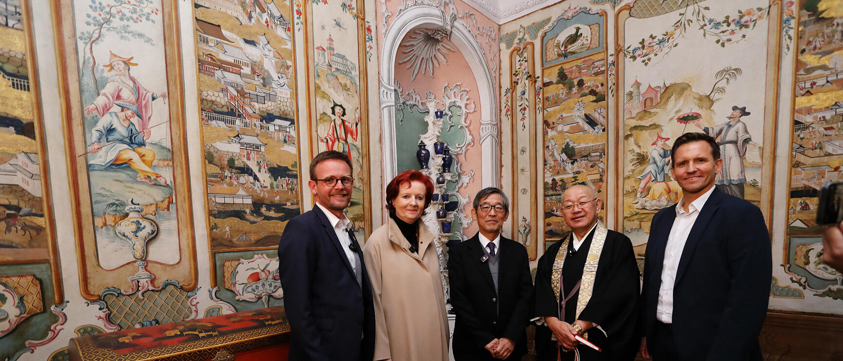 Five people in a room with East Asian wall paintings and lacquered furniture, one person wearing Buddhist robes.