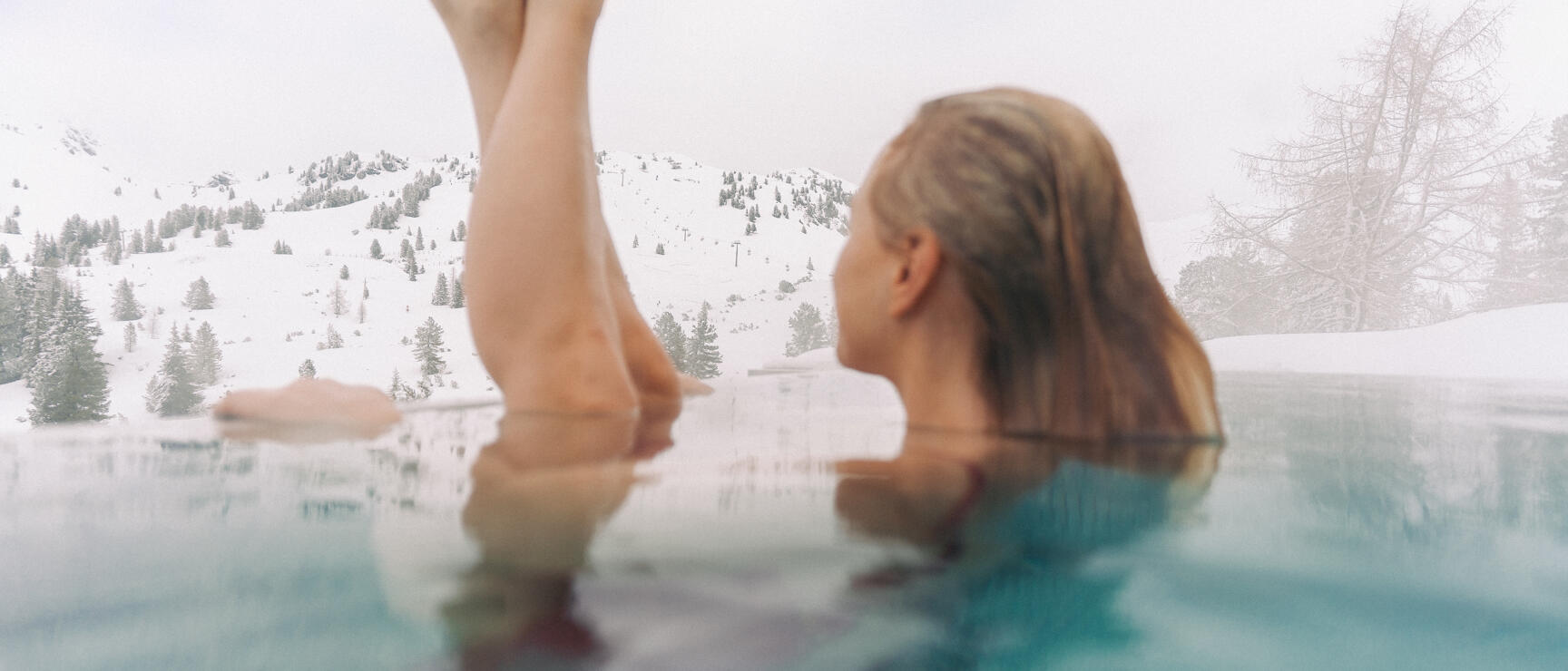 Woman in outdoor pool stretching legs out of the water, snowy mountain landscape with trees in background.