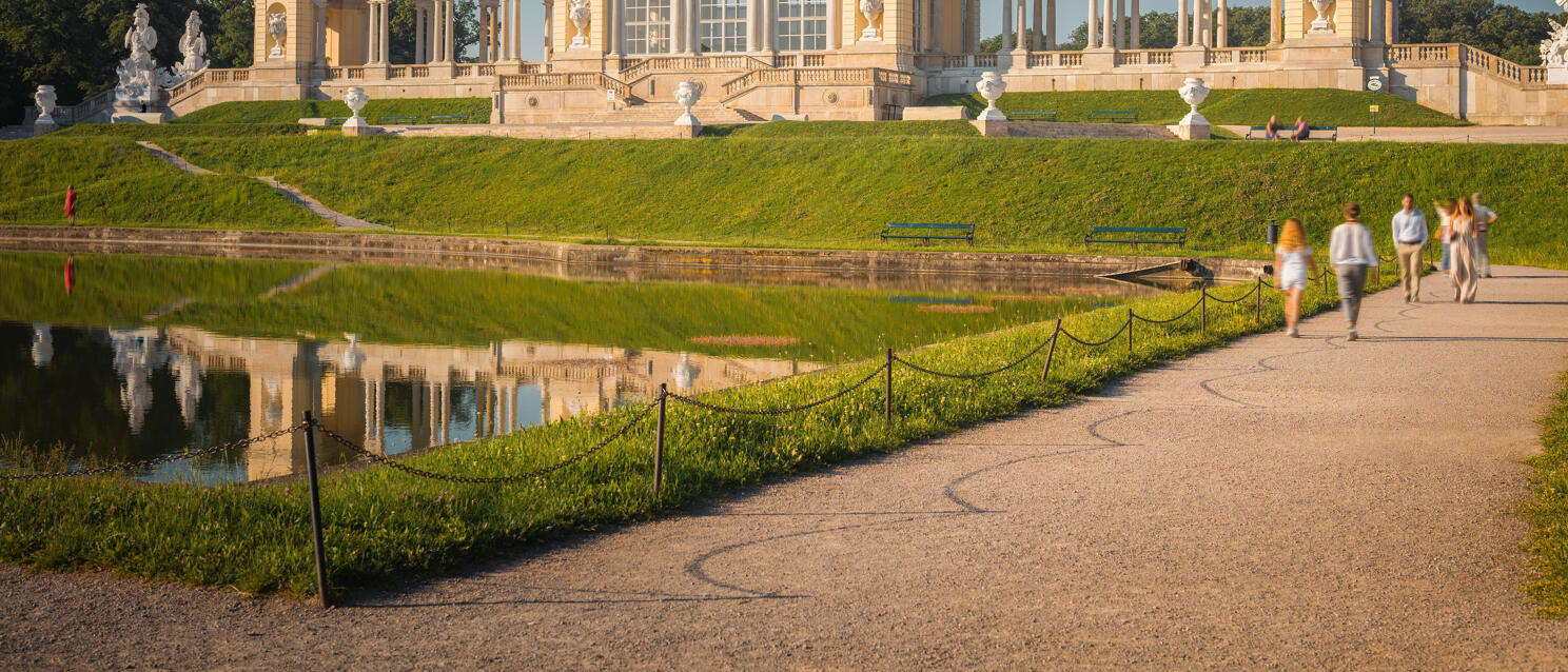 Gloriette in Schönbrunn Palace Park, Vienna, with pond and reflection, strollers on path in front.