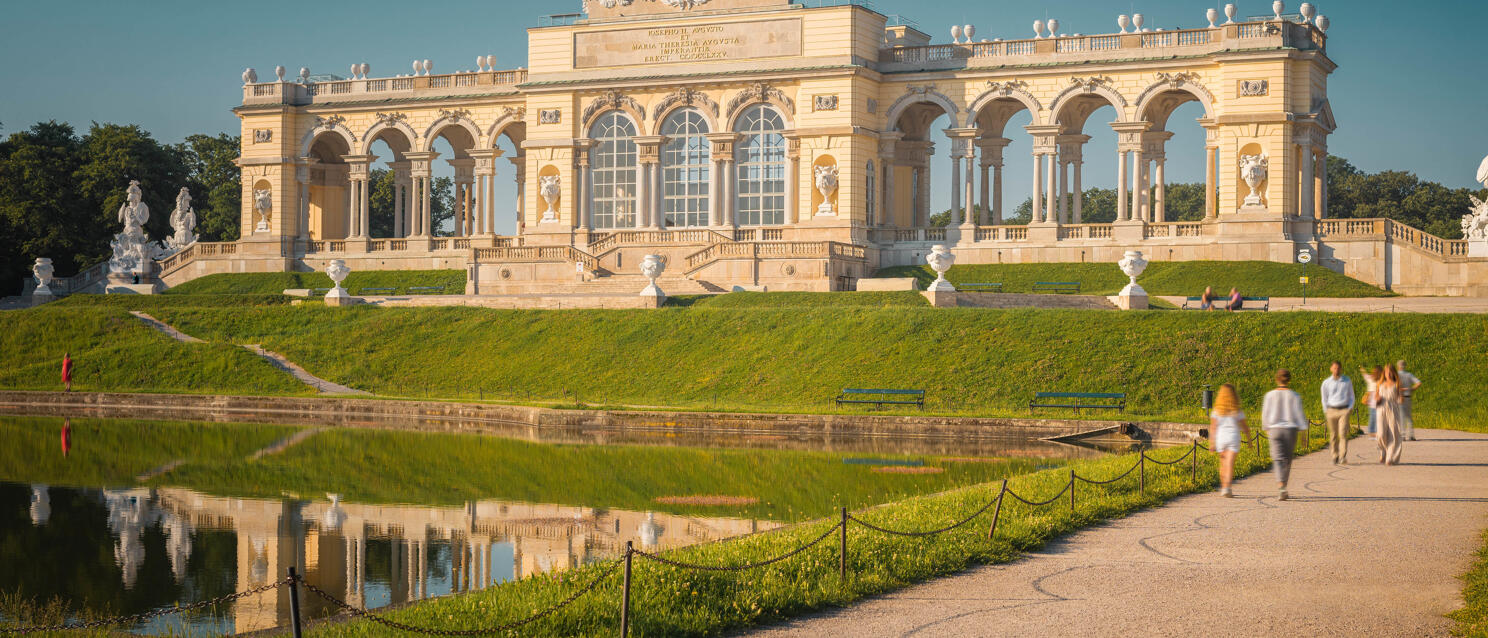 Gloriette in Schönbrunn Palace Park, Vienna, with pond and reflection, strollers on path in front.