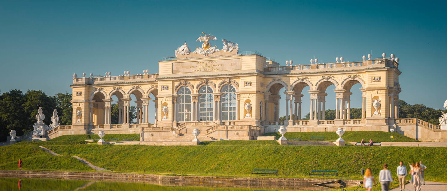 Gloriette im Schlosspark Schönbrunn, Wien, mit Teich und Spiegelung, Spaziergänger auf dem Weg davor.