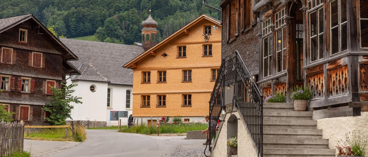 Village square with old wooden houses and church tower, forested mountain slopes in the background, Vorarlberg.