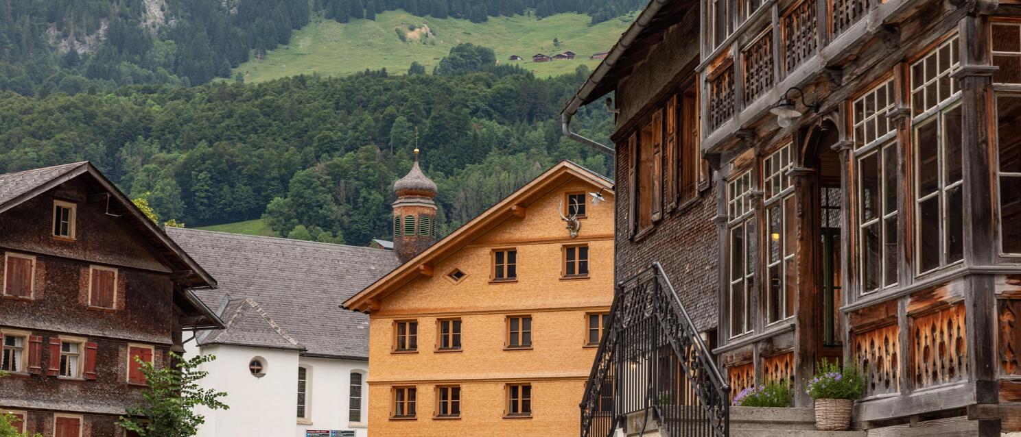 Village square with old wooden houses and church tower, forested mountain slopes in the background, Vorarlberg.