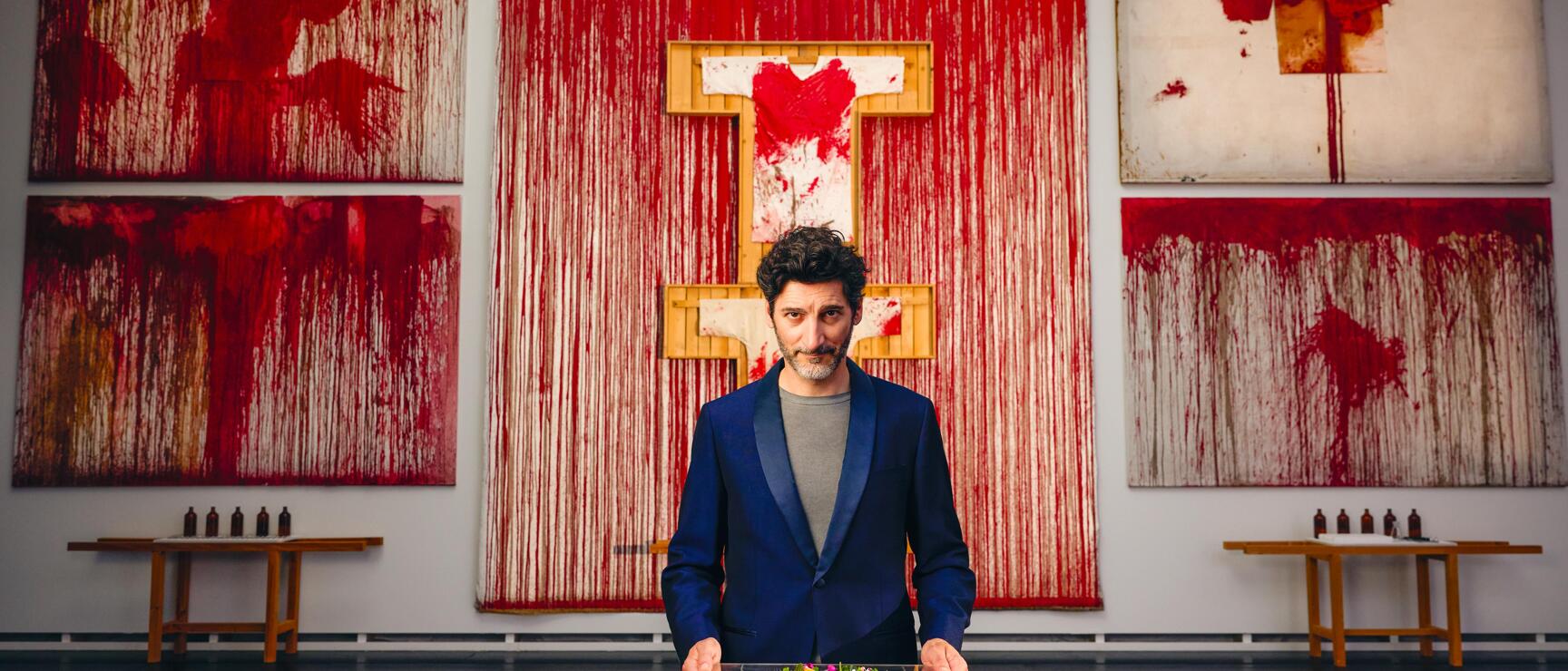 Paul Ivic, Küchenchef des Restaurants Tian, präsentiert ein farbenfrohes Gericht auf einem schwarzen Sockel im Nitsch Museum Mistelbach, im Hintergrund Schüttbilder von Hermann Nitsch in Rot- und Weißtönen.