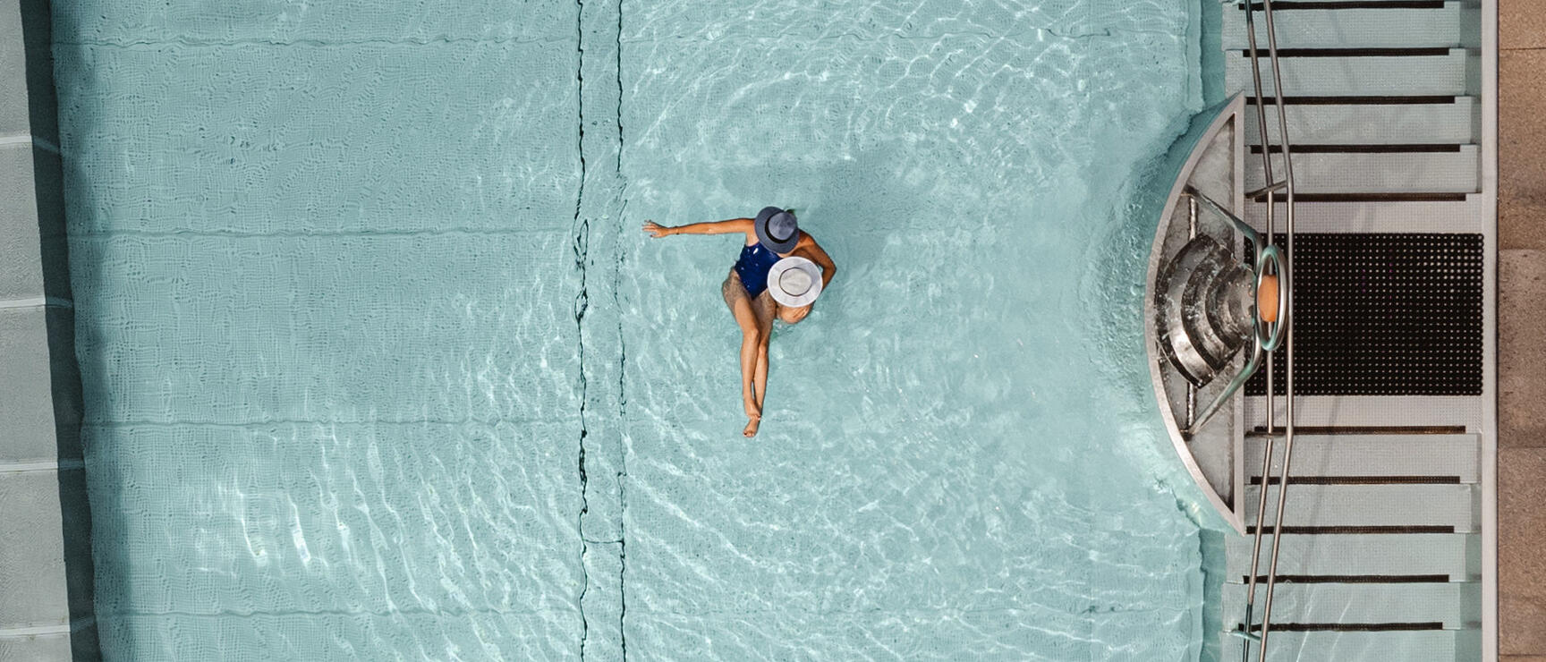 Overhead view: woman in hat wearing turquoise swimsuit floating in blue pool, entry stairs on right.