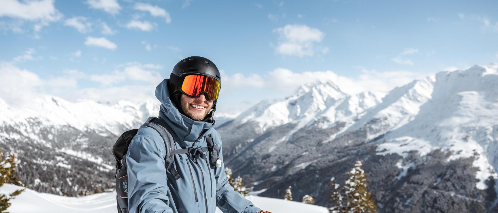 Skier wearing helmet and ski goggles smiling in front of snow-covered mountain peaks under blue sky.