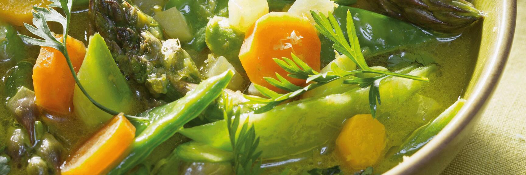 Vegetable soup with green asparagus, carrots, sugar snap peas and herbs in ceramic bowl.