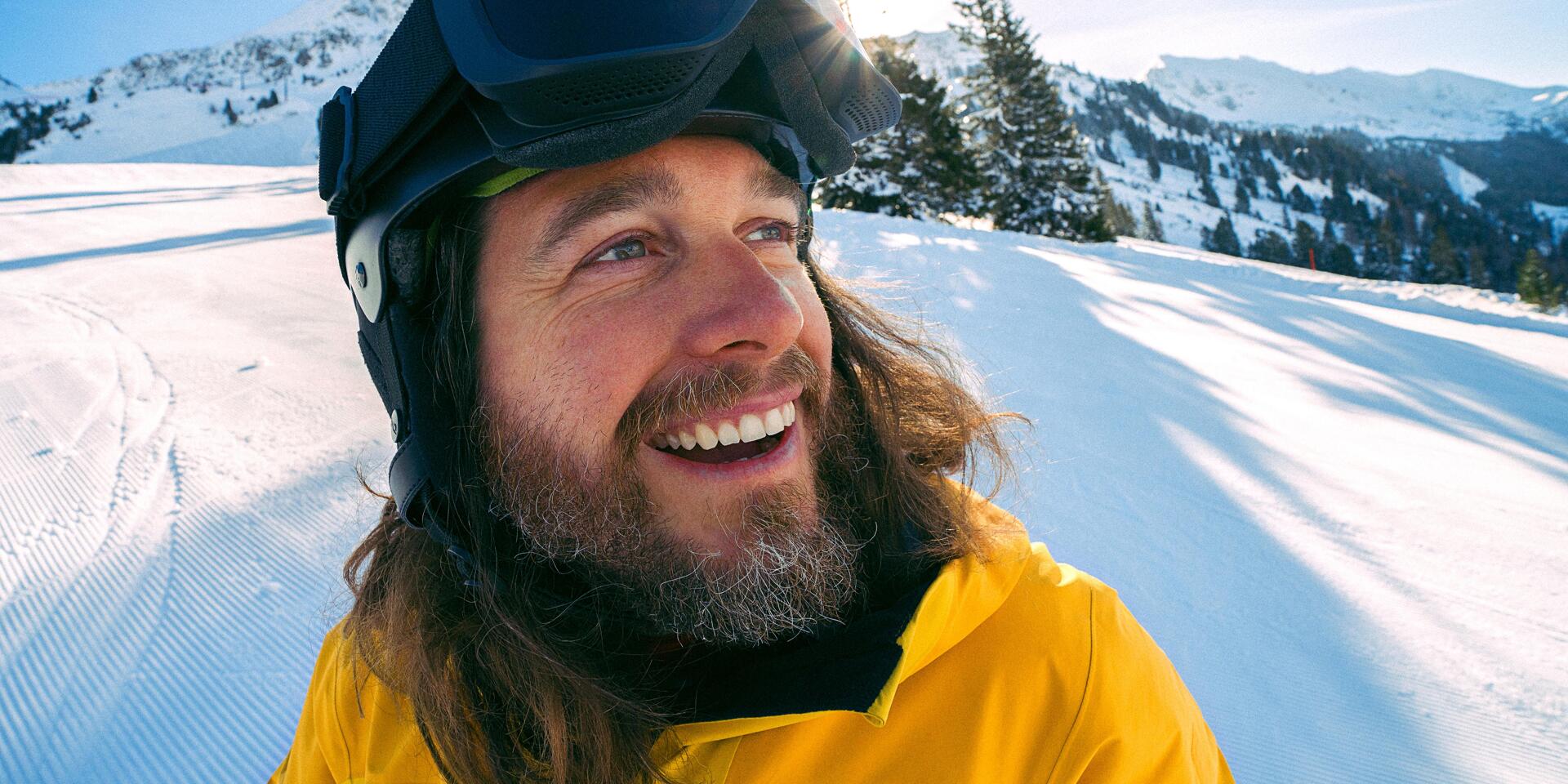 Smiling man wearing ski helmet and yellow jacket on a ski slope, snow-covered mountains in the background.