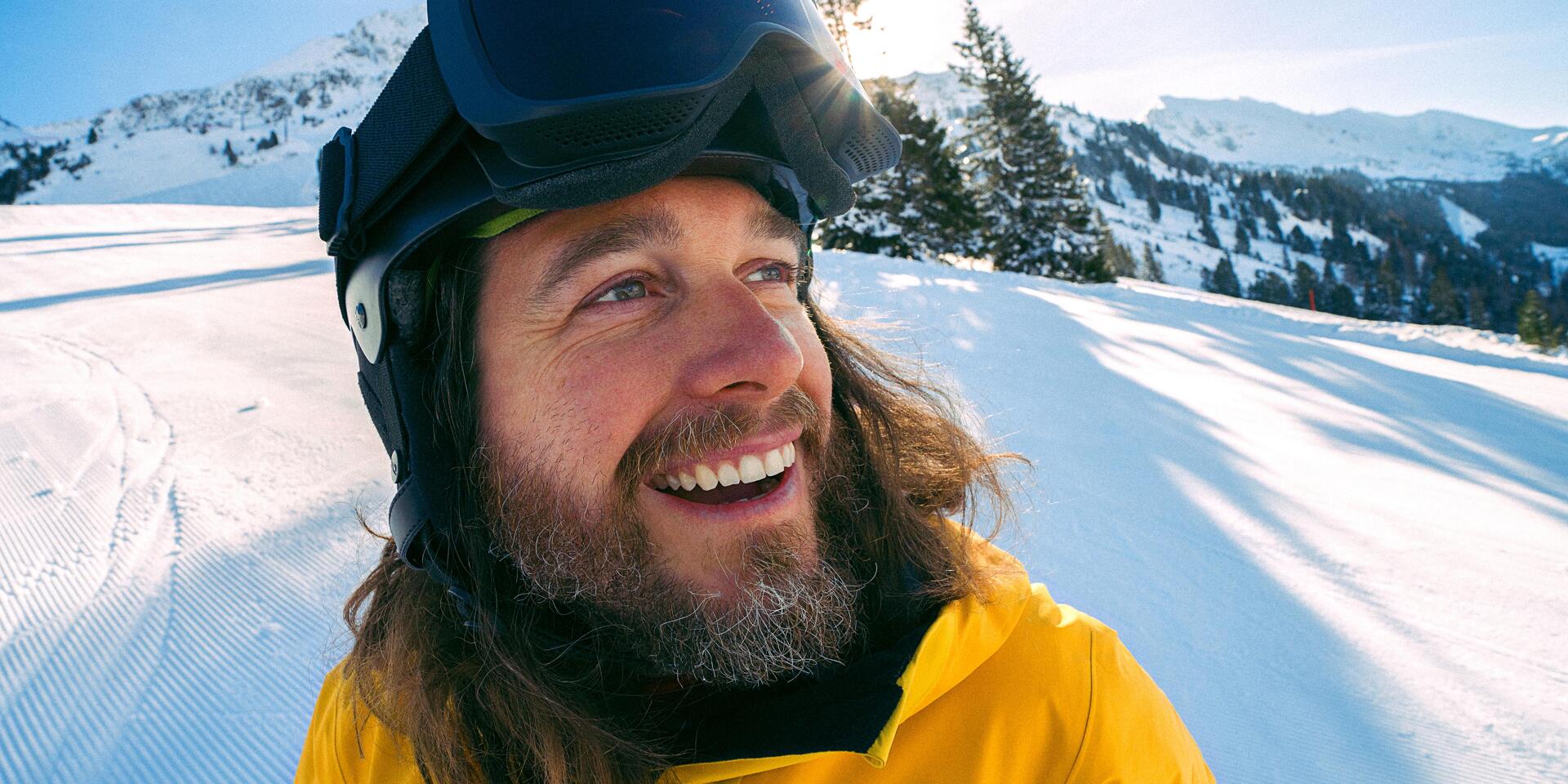 Smiling man wearing ski helmet and yellow jacket on a ski slope, snow-covered mountains in the background.