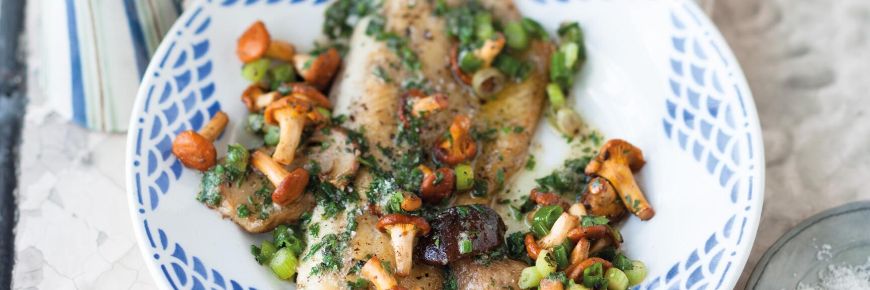 Fried trout with chanterelles, herbs and spring onions on blue and white plate.