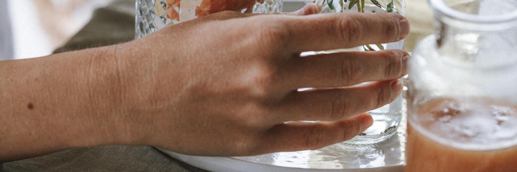 Hands garnishing a glass with ice cubes and fruit with fresh herbs; glasses with syrup nearby.