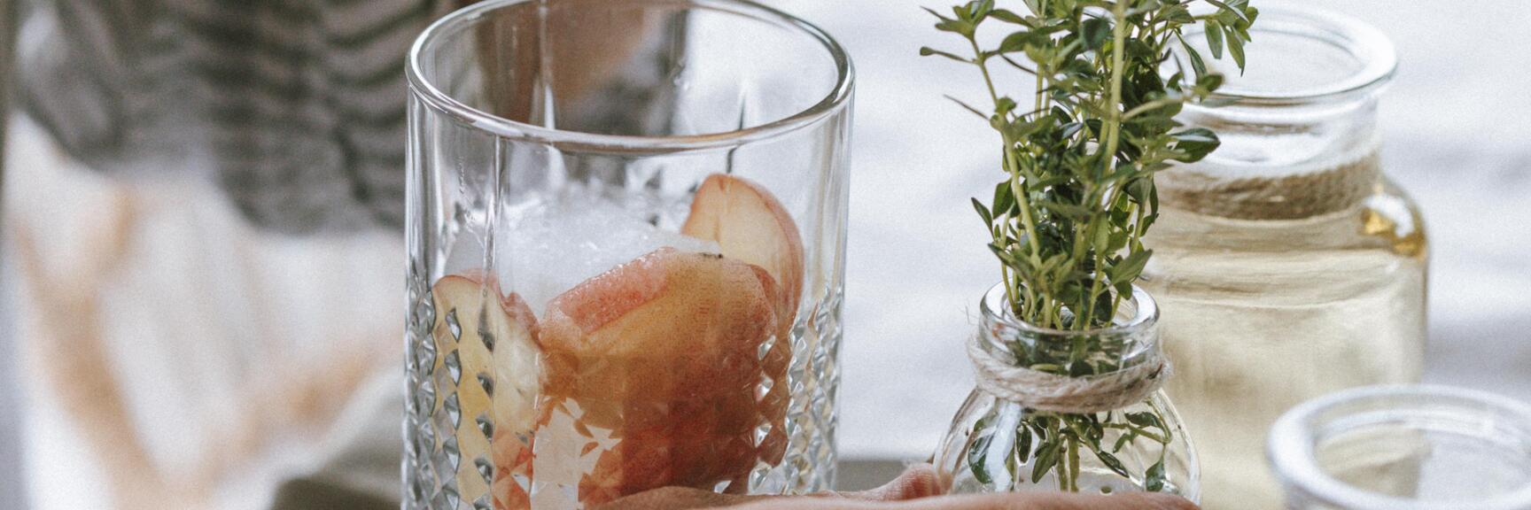 Hands garnishing a glass with ice cubes and fruit with fresh herbs; glasses with syrup nearby.